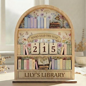 May include: Wooden bookshelf with floral wallpaper, displaying miniature books in pastel shades. The banner reads "BOOKS READ THIS YEAR" above the number "215". The bottom shelf has the text "LILY'S LIBRARY". A small lamp and books are on the top shelf.