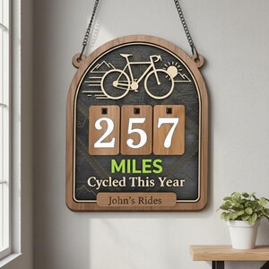 May include: Wooden sign with a bicycle theme and a black chalkboard background. The sign displays "257 MILES Cycled This Year" and "John's Rides." A bicycle and sun graphic are at the top. The sign is suspended by a chain.
