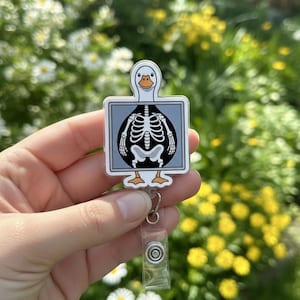 Pode incluir: Um porta-crachá branco e cinza com um ganso de desenho animado com um raio-x de seu esqueleto. O porta-crachá tem uma alça de plástico transparente e um cordão retrátil. O fundo está desfocado com flores verdes e amarelas.