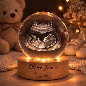 May include: A clear crystal ball with an ultrasound image of a baby inside, illuminated by a warm light. The ball sits on a wooden base engraved with the name "Oliver James", the phrase "Love Begins", and the month and year "June 2026".