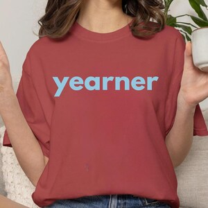 Op de afbeelding: Een baksteenrood T-shirt met korte mouwen en het woord "yearner" in lichtblauwe schreefloze letters op de borst. Het kledingstuk heeft een ronde hals en een relaxte pasvorm, gecombineerd met een blauwe spijkerbroekshort.