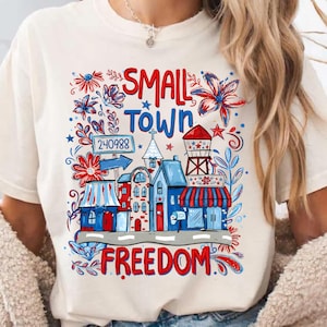 Könnte beinhalten: Ein cremefarbenes Kurzarm-T-Shirt mit einem Grafikdruck, der den Text SMALL TOWN FREEDOM und 240988 enthält. Das Design zeigt rote, weiße und blaue Illustrationen von Gebäuden, einem Wasserturm, Blumen und Sternen im patriotischen Stil.