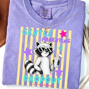 Puede incluir: Una camiseta morada de manga corta con un gráfico de un mapache con gafas de sol que sostiene una bebida con sombrilla. El texto dice GENTLE PARENTING DROPOUT EST THIS MORNING. La camiseta tiene cuello redondo y una etiqueta de la marca Comfort Colors.