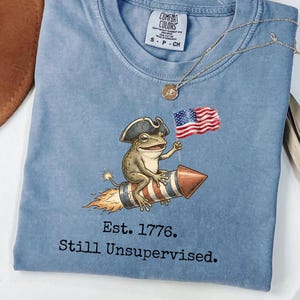 Könnte beinhalten: Hellblaues T-Shirt mit einer Cartoon-Fröschin mit Dreispitz auf einer Rakete, die eine amerikanische Flagge hält. Der Text "Est. 1776. Still Unsupervised." ist darunter gedruckt. Eine goldene Halskette liegt auf dem Shirt.
