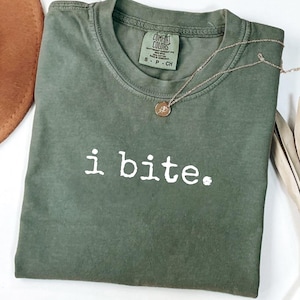 May include: An olive green short-sleeve t-shirt with a crew neckline. The shirt features the white text i bite. printed on the front. A Comfort Colors brand label is visible at the inner neck, indicating a size small and 100% cotton material.