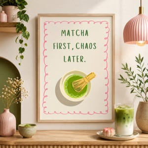 Könnte beinhalten: Ein gerahmter Kunstdruck mit einem rosa gewellten Rand auf cremefarbenem Hintergrund. Der Text MATCHA FIRST, CHAOS LATER ist in Grün gehalten. Darunter befindet sich eine Illustration einer weißen Schale mit grünem Matcha und einem Bambusbesen.