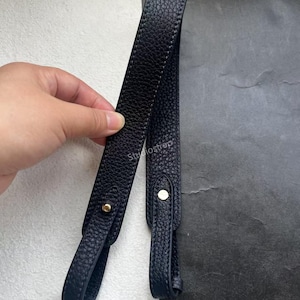 以下が含まれることがあります： ゴールドカラーの金具が付いた、2本のネイビーブルーの革製バッグストラップ。 ストラップは質感のある表面で、ハンドバッグや財布に取り付けるように設計されています。 画像は、白と黒の背景に対してストラップを示しています。