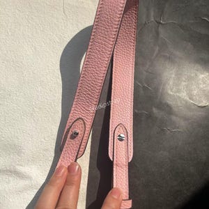 Peut inclure: Une sangle de sac en cuir rose clair avec des fixations argentées. La sangle est en cuir texturé et présente un design simple et élégant. La sangle est présentée sur un fond noir et blanc.