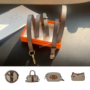 Può includere: Un cinturino per borsa grigio metallizzato con hardware color oro, tra cui una chiusura e una fibbia, è esposto in una scatola arancione. Il cinturino è mostrato arrotolato e srotolato. Sotto ci sono quattro piccole borse con il logo Gucci.