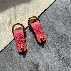 May include: Two pink leather key fobs with gold-colored hardware. Each fob has a D-ring and a gold-colored stud. The fobs are set against a textured gray surface and a cream-colored surface.