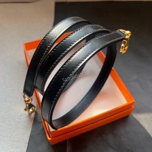 Può includere: Cinturino in pelle nera con finiture dorate, arrotolato all'interno di una scatola arancione. Il cinturino è liscio e sembra nuovo. Il testo "Studiostrap" è visibile sul cinturino.