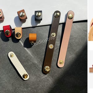 May include: A collection of leather bag accessories in various colors, including brown, red, and cream. The accessories feature gold-colored hardware, such as rivets and snaps. The image also includes a marble coaster and the text "Studiostrap".