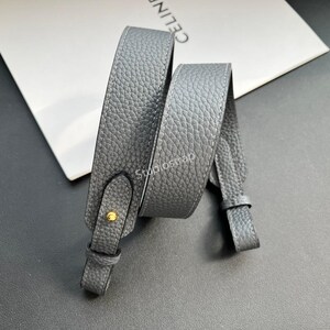 以下が含まれることがあります： グレーの革製バッグストラップ。表面はテクスチャード加工。ストラップには金色の金属ボタンと、取り付け用のループが両端に付いています。「CELINE」の文字が白い背景に部分的に見えます。