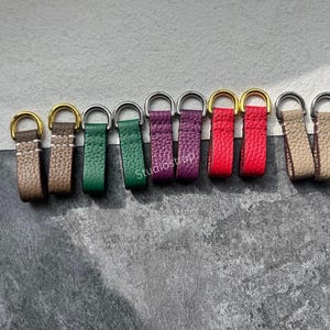 May include: A collection of colorful leather zipper pulls with metal rings. The pulls come in various colors, including brown, green, purple, red, and tan. The metal rings are either gold or silver in color. The text "Studiostrap" is visible on some of the pulls.