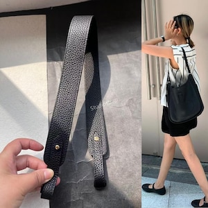 Op de afbeelding: Zwarte leren schouderriem met goudkleurige hardware. De riem wordt getoond in een close-up, en ook op een zwarte leren handtas. De handtas wordt gedragen door een persoon die een zwart-wit gestreepte top en een zwarte short draagt.