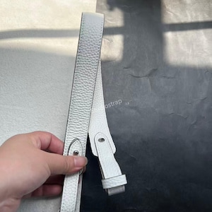 以下が含まれることがあります： 白の革製バッグストラップ。表面は質感があり、金属製のバックルと小さなループが付いています。ストラップは白黒の背景に表示されています。
