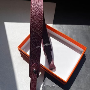 May include: A burgundy leather strap with a textured finish, likely for a bag or accessory. The strap is displayed against a white and black backdrop, with an orange and white box in the background. The text "Studio Straps" is visible.