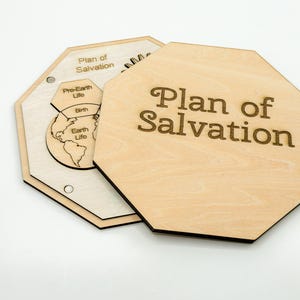 May include: Wooden octagonal plaque with the words "Plan of Salvation" engraved on the front. The plaque is light brown with a darker brown border. Another plaque shows a diagram of the plan.