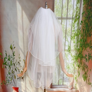 May include: A white bridal veil with a scalloped edge is displayed on a mannequin. The veil is made of a sheer fabric and has a delicate, layered design. The mannequin is on a stand in front of a window with greenery.