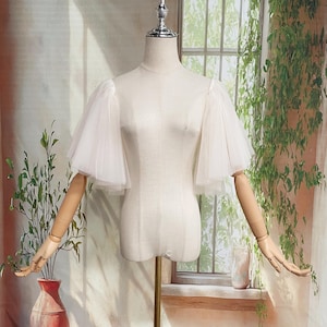 May include: A cream-colored dress form with articulated wooden arms and a metal stand. The dress form has sheer, puffy sleeves. The background features a window with greenery and a red vase.