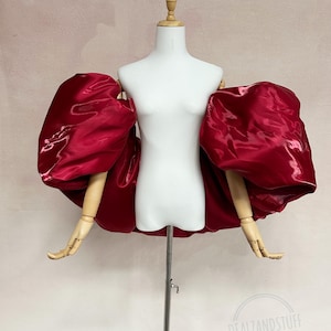 May include: A deep red, off-the-shoulder garment featuring voluminous, oversized puff sleeves made of a shiny, satin-like fabric. The piece is displayed on a white mannequin torso with articulated wooden arms and includes the text DEALZANDSTUFF.