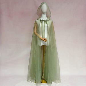 May include: A floor-length, sage green sheer organza cloak featuring a hood and a delicate ribbon tie closure at the neckline. The garment is displayed on a mannequin, showcasing its translucent, flowing fabric and layered construction.