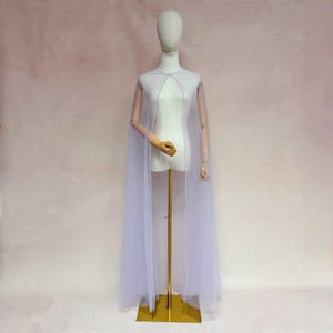 May include: A floor-length, sheer white cape made of lightweight tulle fabric. The cape features a simple, open-front design that drapes from the shoulders, creating a flowing silhouette. It is displayed on a neutral-toned mannequin stand.