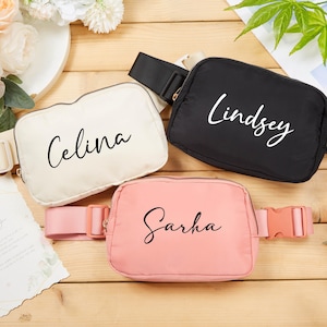 Może przedstawiać: Trzy spersonalizowane nerki w kolorze kremowym, czarnym i różowym. Każda nerka ma imię w skrypcie kursywnym: Celina, Lindsey i Sarha. Nerki mają regulowane paski i plastikowe klamry. Tło zawiera kwiaty i drewnianą powierzchnię.