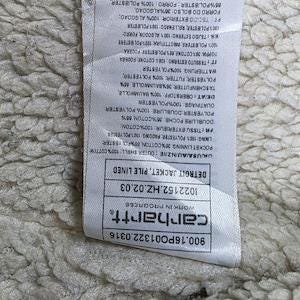 Op de afbeelding: Close-up van een Carhartt-label met tekst waaronder "DETROIT JACKET, PILE LINED" en het Carhartt-logo. Het label is wit met zwarte tekst en bevestigd aan een getextureerde, gebroken witte stof.