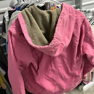 Op de afbeelding: Een roze jas met capuchon en een beige sherpa voering hangt aan een witte hanger. De jas heeft een volledige ritssluiting en een capuchon met trekkoord. De jas is gemaakt van een zacht materiaal en lijkt in goede staat te zijn.