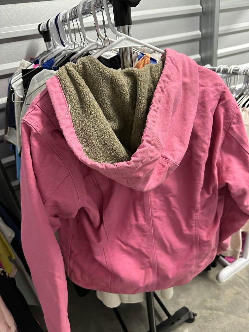 Op de afbeelding: Een roze jas met capuchon en een beige sherpa voering hangt aan een witte hanger. De jas heeft een volledige ritssluiting en een capuchon met trekkoord. De jas is gemaakt van een zacht materiaal en lijkt in goede staat te zijn.