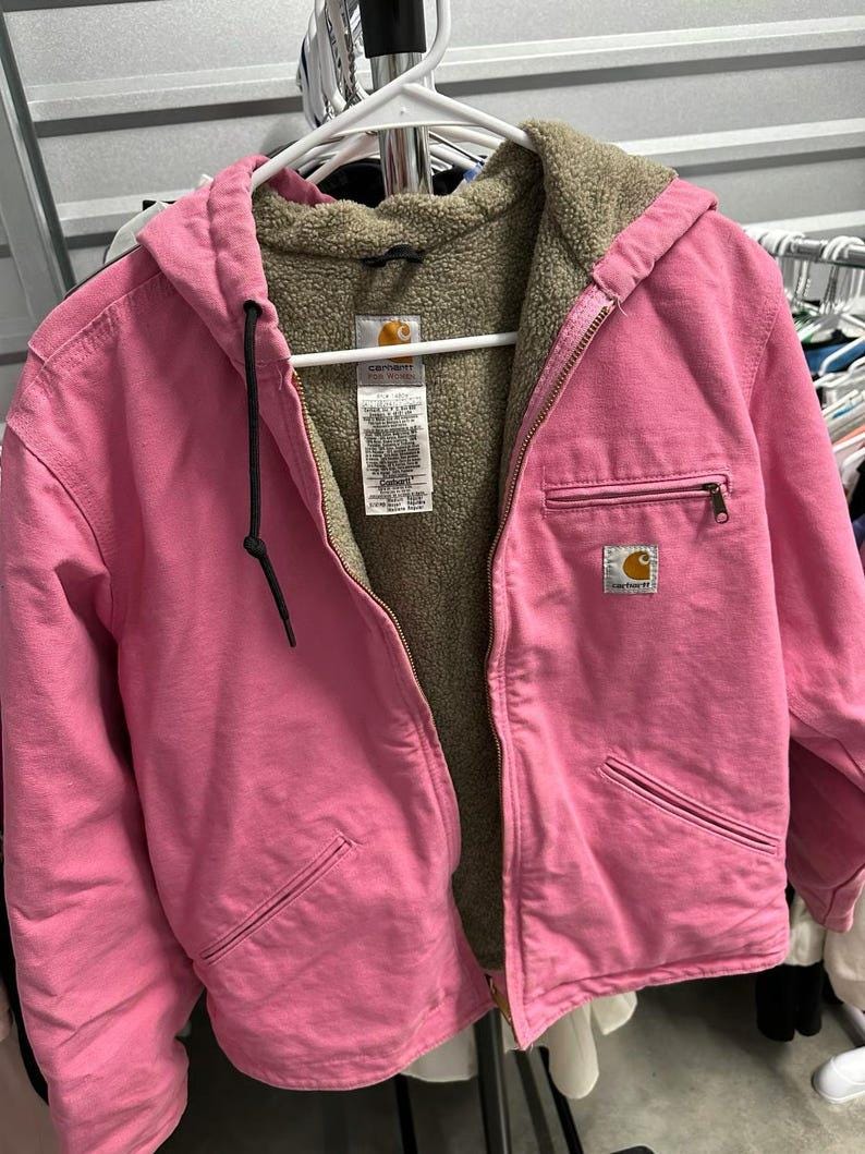Op de afbeelding: Een roze Carhartt jas met capuchon en een bruine, fuzzy binnenkant. De jas heeft een ritssluiting, twee voorzakken en een kleine Carhartt-logopatch. De capuchon heeft een zwart trekkoord. De jas hangt aan een witte hanger.