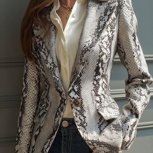 May include: A snakeskin-patterned blazer in shades of gray and white, featuring a notched lapel, two front pockets, and button closures. The blazer is worn over a white collared shirt and dark blue jeans. A gold necklace is visible.