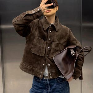 May include: A brown suede jacket with silver buttons and two front pockets. The jacket is paired with blue jeans and a dark brown leather bag. A silver belt with a decorative buckle is also visible.