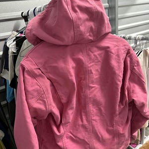 Op de afbeelding: Een roze jas met capuchon en een volledige ritssluiting. De jas heeft een grote capuchon en een iets oversized pasvorm. De achterkant van de jas is zichtbaar, met details van de stiksels en de textuur van de stof. De jas hangt aan een rek met andere kleding.