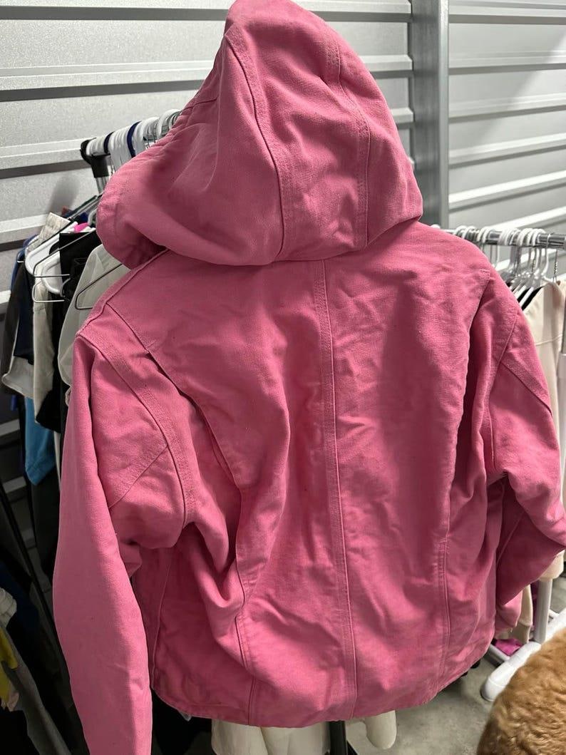 Op de afbeelding: Een roze jas met capuchon en een volledige ritssluiting. De jas heeft een grote capuchon en een iets oversized pasvorm. De achterkant van de jas is zichtbaar, met details van de stiksels en de textuur van de stof. De jas hangt aan een rek met andere kleding.