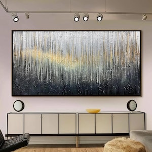 May include: Large abstract painting with a black frame. The artwork features vertical lines in silver, gold, and black, creating a textured effect. The painting hangs above a white cabinet with black accents, complemented by a black leather chair and a fluffy ottoman.