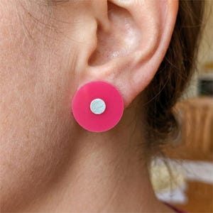 May include: A circular stud earring featuring a vibrant hot pink disc with a smaller, centered silver-toned metallic circle. The earring is positioned on an earlobe, showcasing a flat, geometric design with a smooth, matte finish on the pink surface.