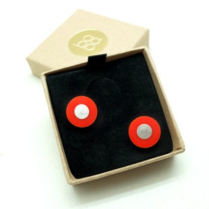 May include: A pair of red and silver stud earrings in a small brown box. The earrings are circular with a silver center and a red outer ring.