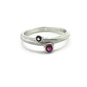 Peut inclure: Une bague en argent avec deux petites pierres précieuses. La plus grande pierre est un rubis rose vif, et la plus petite est une améthyste violette foncée.