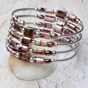 May include: A silver-coloured, multi-strand bracelet featuring rectangular and oval beads in shades of mauve and silver. The bracelet is coiled and rests on a white seashell, showcasing a unique jewellery design.