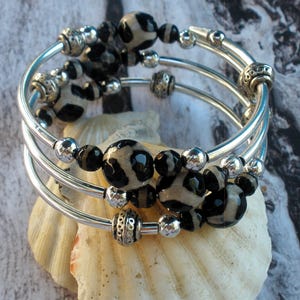 May include: A silver bangle bracelet with three rows of black and white agate beads. The bracelet is on a white seashell.