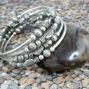 May include: A set of silver bangles with decorative beads and leaf accents. The bangles are stacked on top of each other and are resting on a gray stone.