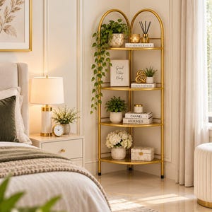 Op de afbeelding: Een hoekrek met vier niveaus van goudkleurig metaal met gebogen bovenkanten. De planken bevatten diverse decoratieve items, waaronder potplanten, een ingelijst bord met de tekst Good Vibes Only, een kaars en boeken met de titels Kinfolk, Louis Vuitton, Chanel en Hermes.