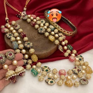 Puede incluir: Un conjunto de collar y pendientes ornamentado con perlas de imitación, piedras preciosas de colores y detalles metálicos dorados. El collar tiene múltiples hilos con elementos decorativos. Los pendientes tienen un diseño a juego. También hay una pulsera dorada.