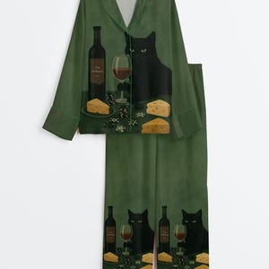 May include: A dark green pajama set featuring a button-down long-sleeve shirt and matching pants. The fabric displays a repeating graphic of a black cat sitting at a table with a wine bottle, a glass of red wine, cheese wedges, and poker chips.
