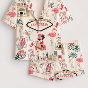 May include: Cream-colored pajama set with a Las Vegas theme. The short-sleeved top and shorts feature a print of flamingos, dice, playing cards, and the "Welcome to Fabulous Las Vegas" sign. The set has pink trim.