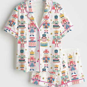 May include: A white pajama set featuring a short-sleeved button-down shirt and matching shorts. The fabric is covered in a repeating pattern of colorful nutcracker figures, drums, and gold stars, accented with pink piping along the edges.