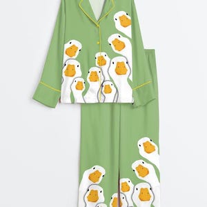 以下が含まれることがあります： ライトグリーンのパジャマセット。長袖のボタンダウンシャツと襟付きのワイドパンツで構成されています。生地には、オレンジ色のくちばしを持つ白いガチョウの繰り返し模様が描かれており、縁には黄色のパイピングが施されています。