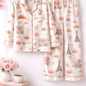 May include: Cream-coloured pyjama set with a Parisian theme. The set includes a button-down shirt and trousers, both decorated with illustrations of the Eiffel Tower, croissants, macarons, and floral accents. The trim is a reddish-brown colour.